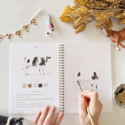 Open book with art supplies and a cow illustration on a white surface