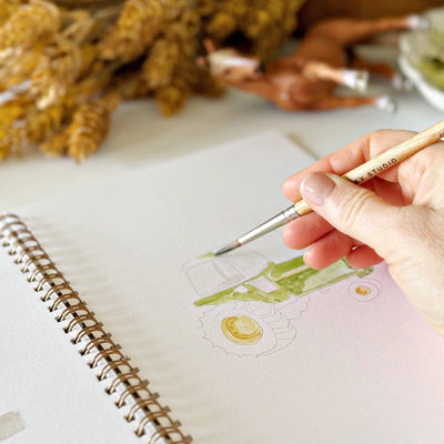 Person painting a design on a notebook with a paintbrush, surrounded by dried plants.