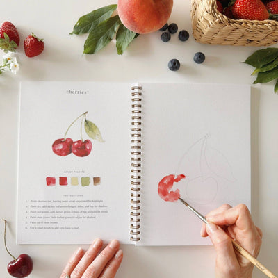 Person painting cherries in a sketchbook with fruits and berries on a table