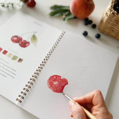 Person painting a cherry on a sketchbook with fruits in the background