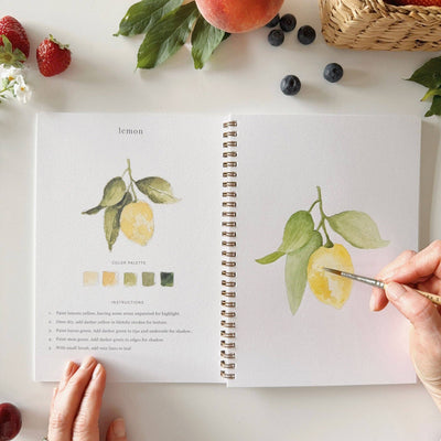 Artbook open to pages with lemon illustrations and color palettes, surrounded by fruits on a white surface.