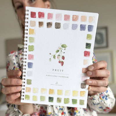 Person holding a watercolor workbook with color swatches and 'Fruit' text.