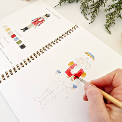 Person coloring a nutcracker illustration in a sketchbook with a color swatch guide.