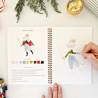 Person coloring a mouse king illustration in a coloring book with a color palette on a white surface.