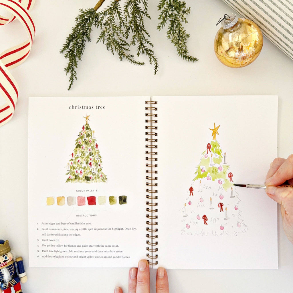 Open book with Christmas tree illustrations and color palette on a white surface with holiday decorations.