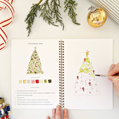 Open book with Christmas tree illustrations and color palette on a white surface with holiday decorations.