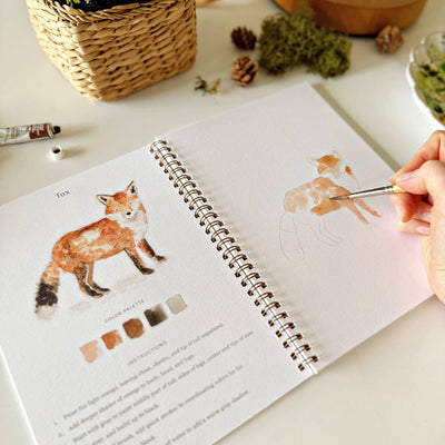 Person painting a fox illustration in a sketchbook with art supplies on a table.