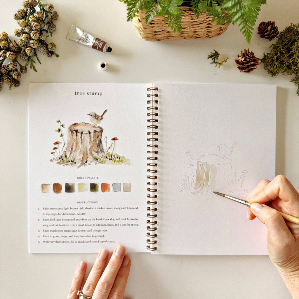 Open book with nature-themed illustrations and color palette on a desk with decorative items.