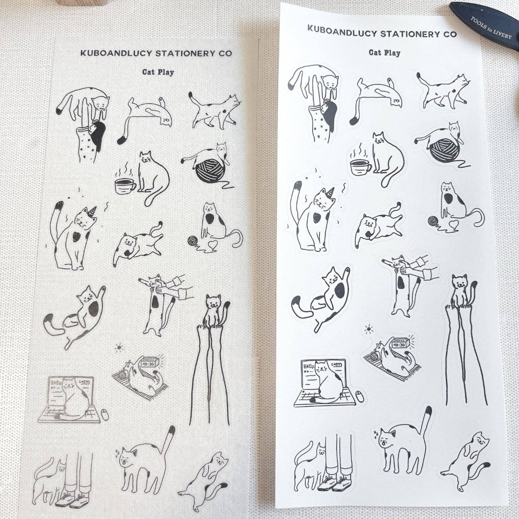 Two sheets of black and white cat illustrations by Kuboandlucy Stationery Co. on a white surface with a wooden stamp set in the background.