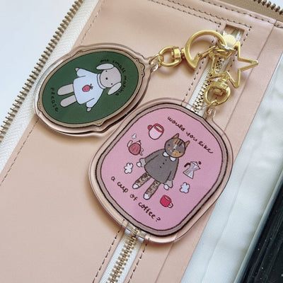Two keychains from Kuboandlucy with cartoon designs on a beige pouch, available at Lighthouse Paper Co. in Melbourne, FL