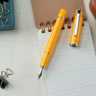 Two yellow pens on a notebook with office supplies in the background.
