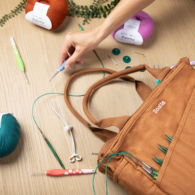 Person working with knitting tools and yarn on a wooden surface, with a brown bag labeled 'Soolia' containing needles.