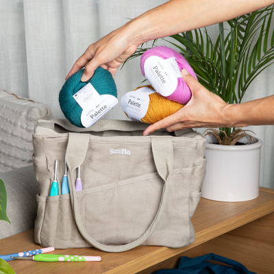 Person placing yarn balls into a beige bag on a wooden surface with plants in the background