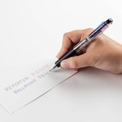 Hand holding a three-color ballpoint pen writing on paper with 'REPORTER 3-COLOR BALLPOINT PEN' text.